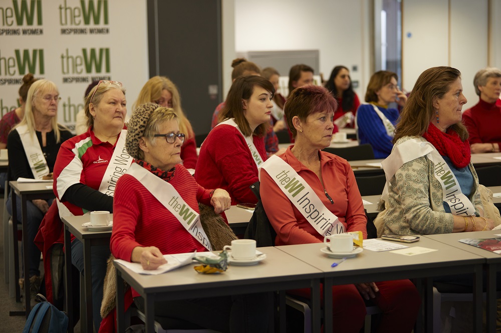 A group of women wearing WI banners sitting at tables at a meeting
