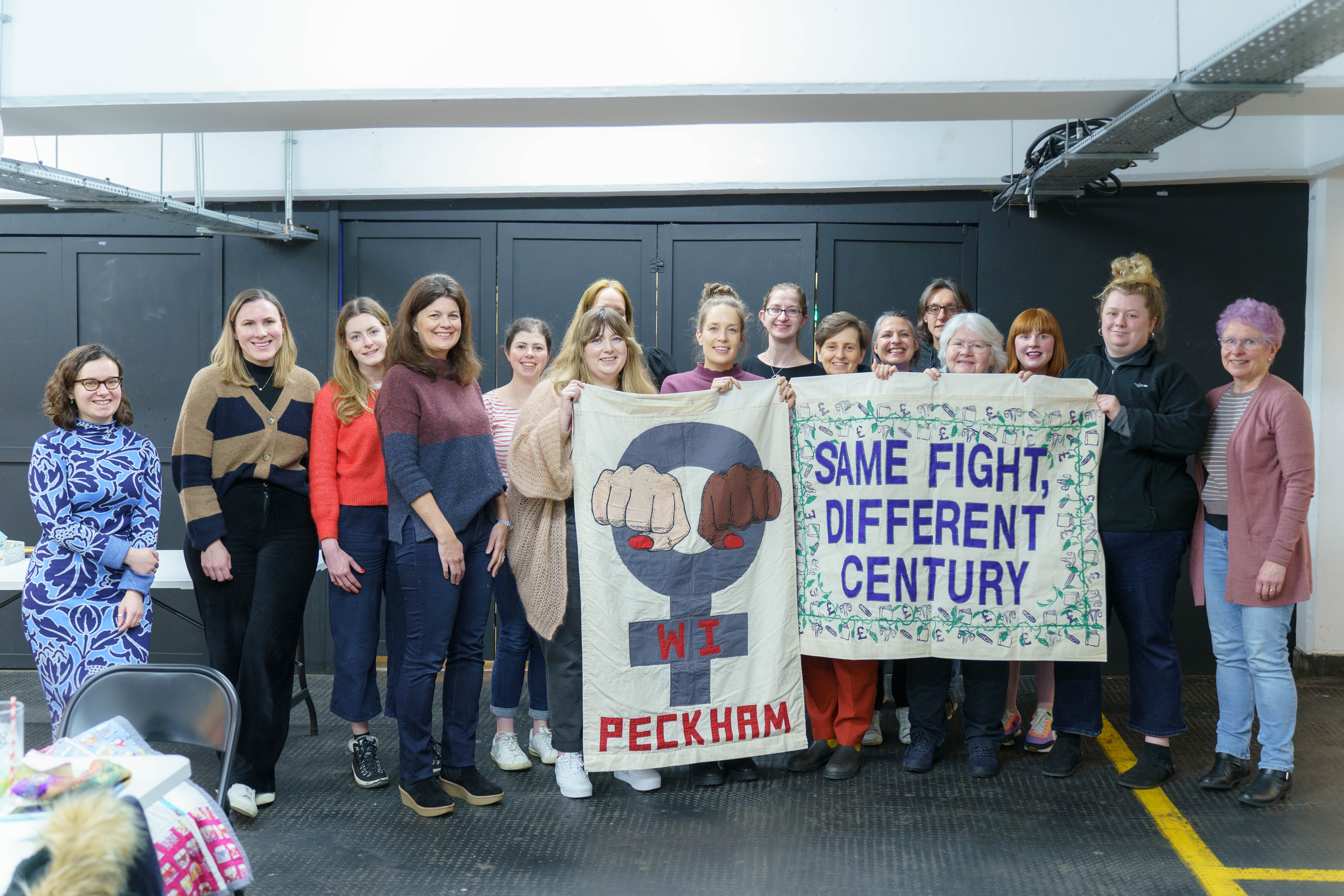 A group of women standing in a room holding two signs. The sign on the left shows the female gender symbol. The sign on the right reads 'same fight different century'.