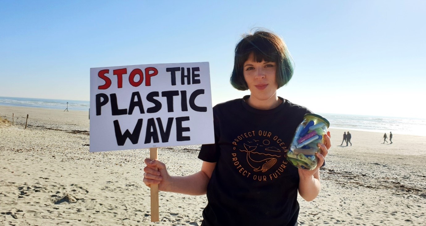 Picture of Ella Daish on the beach holding a sign that reads 'Stop the plastic wave