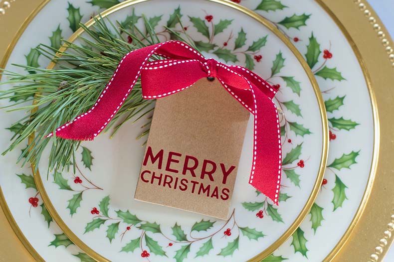 Brown tag tied with red ribbon and the message 'Merry Christmas' on top of a sprig of holly on a plate decorated with holly