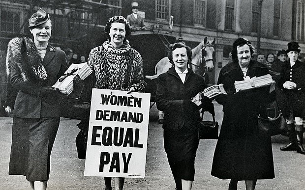 Four women next to each other with one holding up a sign saying "Women demand equal pay"