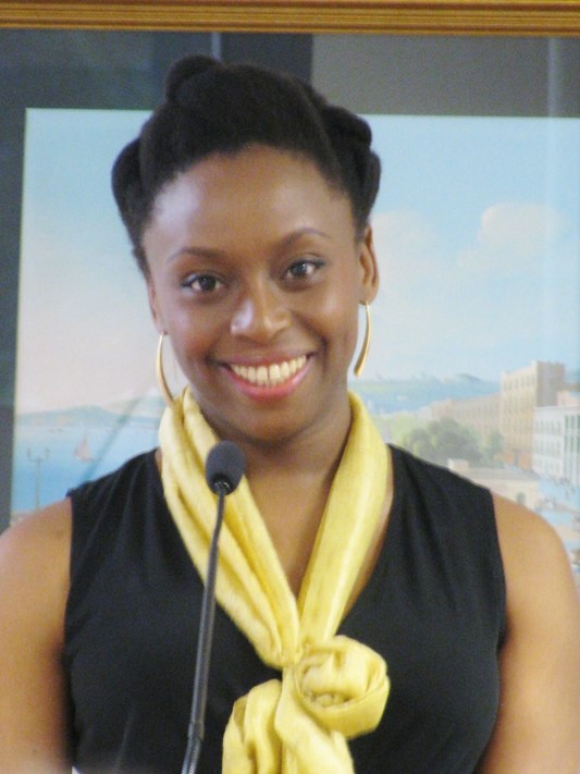 Chimamanda Ngozi Adichie standing behind a microphone smiling towards the camera